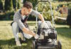 Man cutting grass with lawn mover in the back yard. Male in a black apron. Guy repairs.